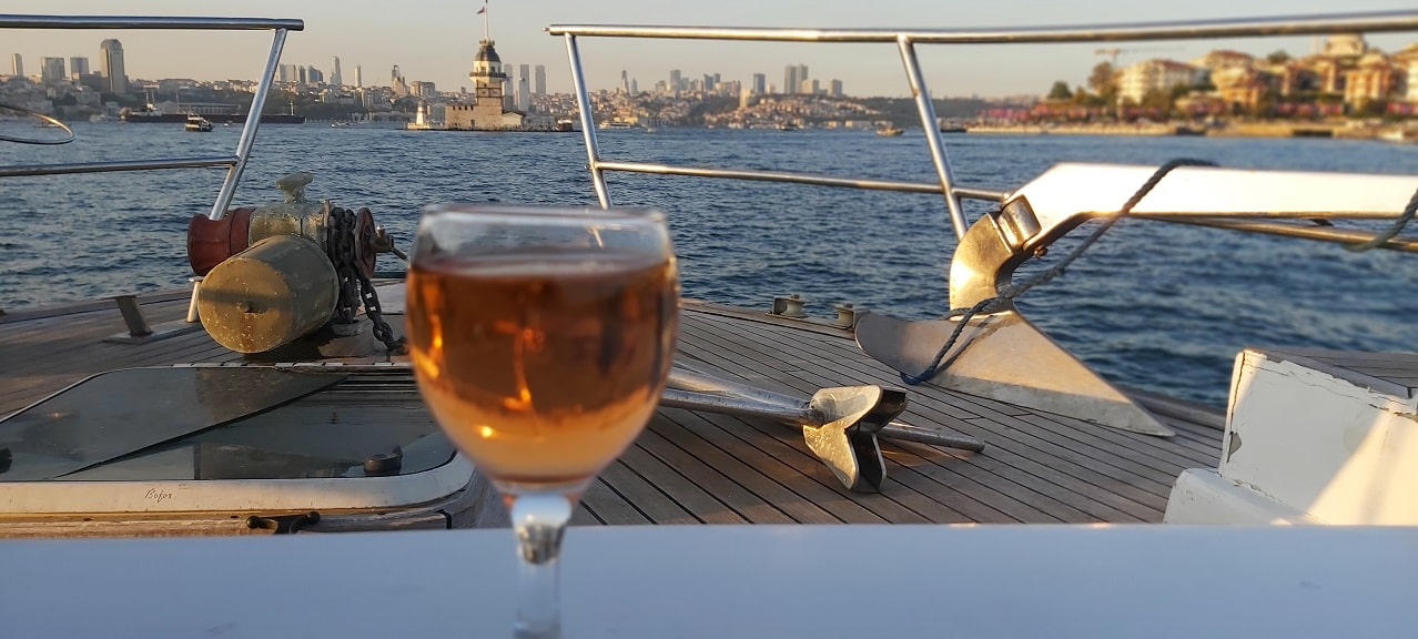A glass of red wine on a wooden table on a boat in the Bosphorus, with the sun shining in the background.